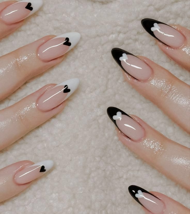 Almond nails with a glossy nude pink base, alternating black and white French tips, and small contrasting heart nail art accents.