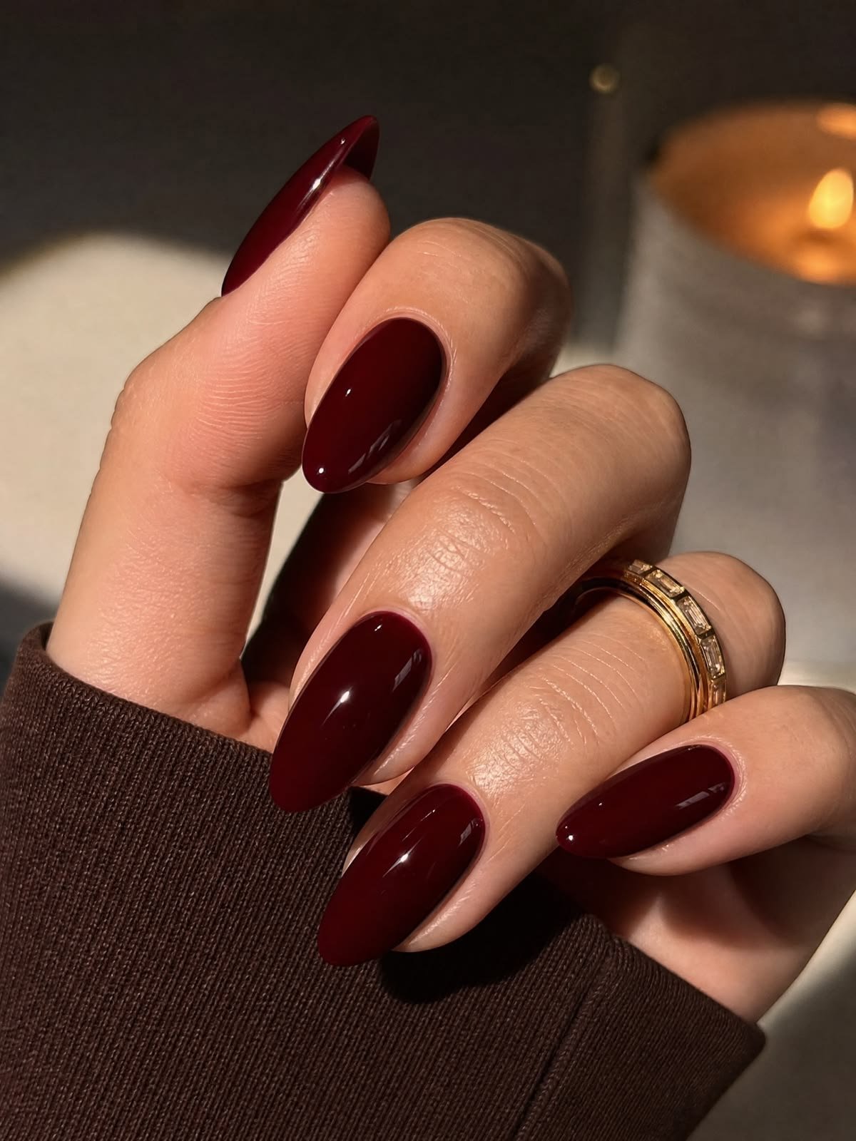 Close-up of a hand with long almond-shaped dark burgundy glossy nails wearing a thin gold ring against a cozy background.