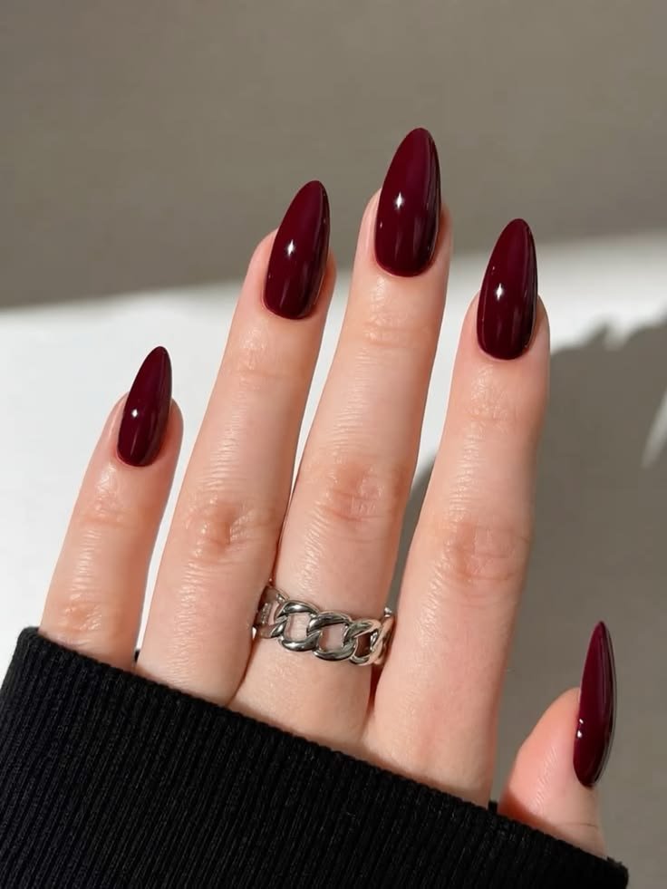 Close-up of almond-shaped glossy dark burgundy nails wearing a chunky silver chain ring against a dark sleeve background.