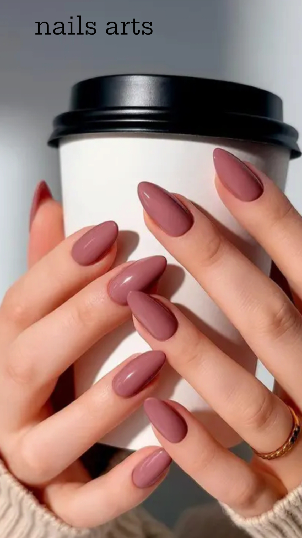 Close-up of hands holding a white coffee cup showcasing glossy dusty rose almond nails and a gold ring.